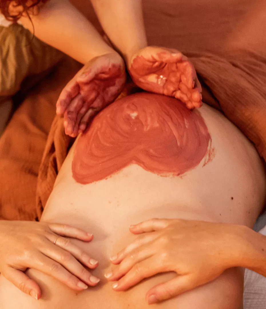 La photo a été prise de ciel. On voit le ventre d'une femme enceinte allongée, recouvert d'un cataplasme de plantes et argiles en forme de coeur.